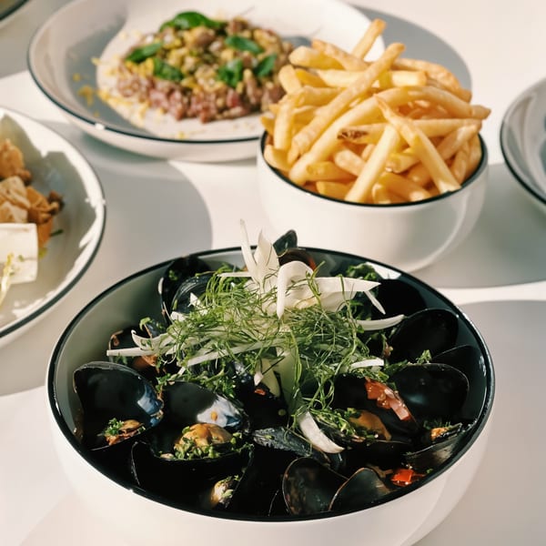 A bowl of mussels with herbs and fennel, alongside fries and another dish, on a white table.